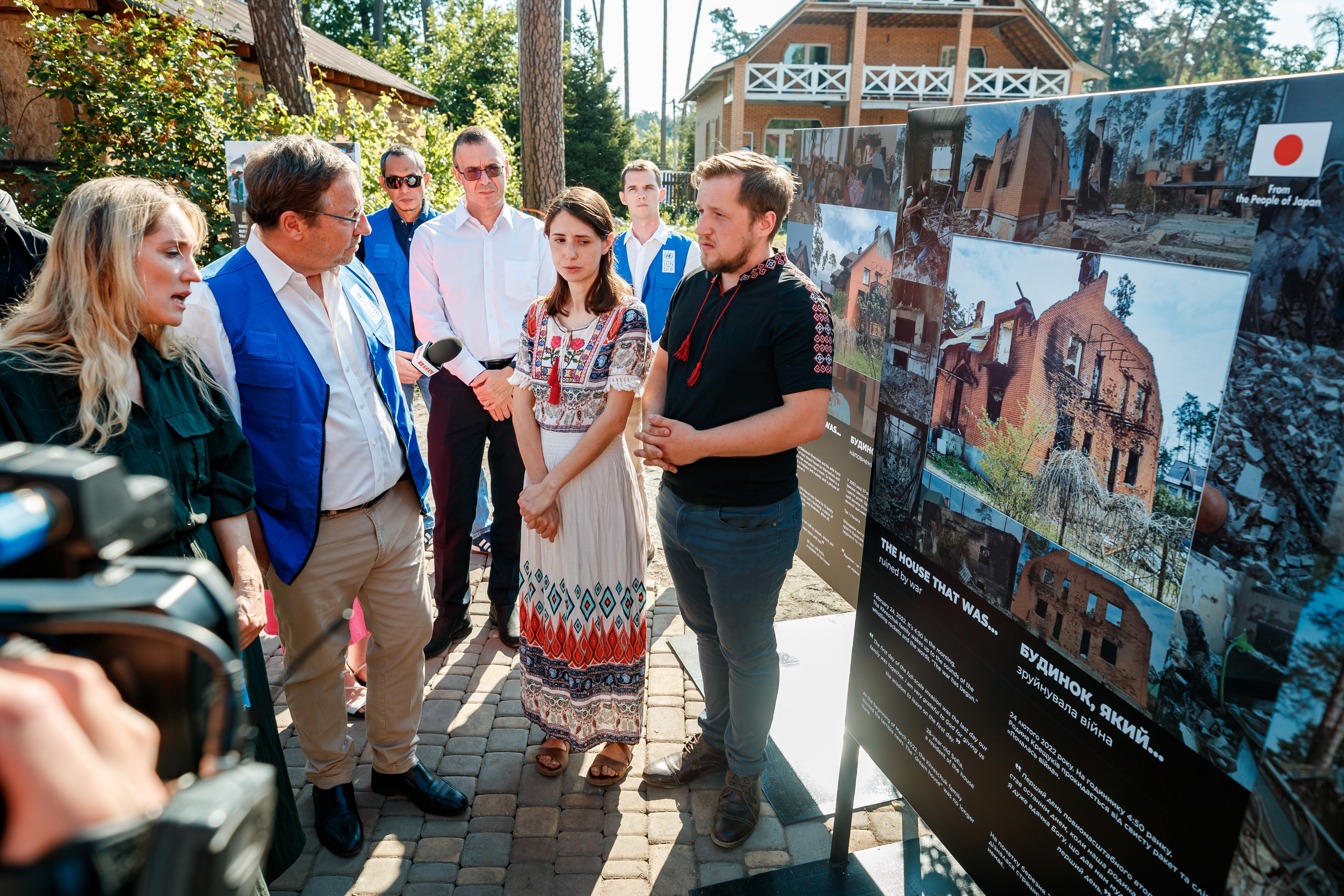 UNDP-Leiter Achim Steiner steht umringt von einer Dolmetscherin, UNDP-Mitarbeitenden vor zwei ukrainischen Personen. Im Hintergrund ihr wiederaufgebautes Haus und davor eine Erklärtafel auf der das vom Ukrainekrieg zerstörte Haus abgebildet ist.