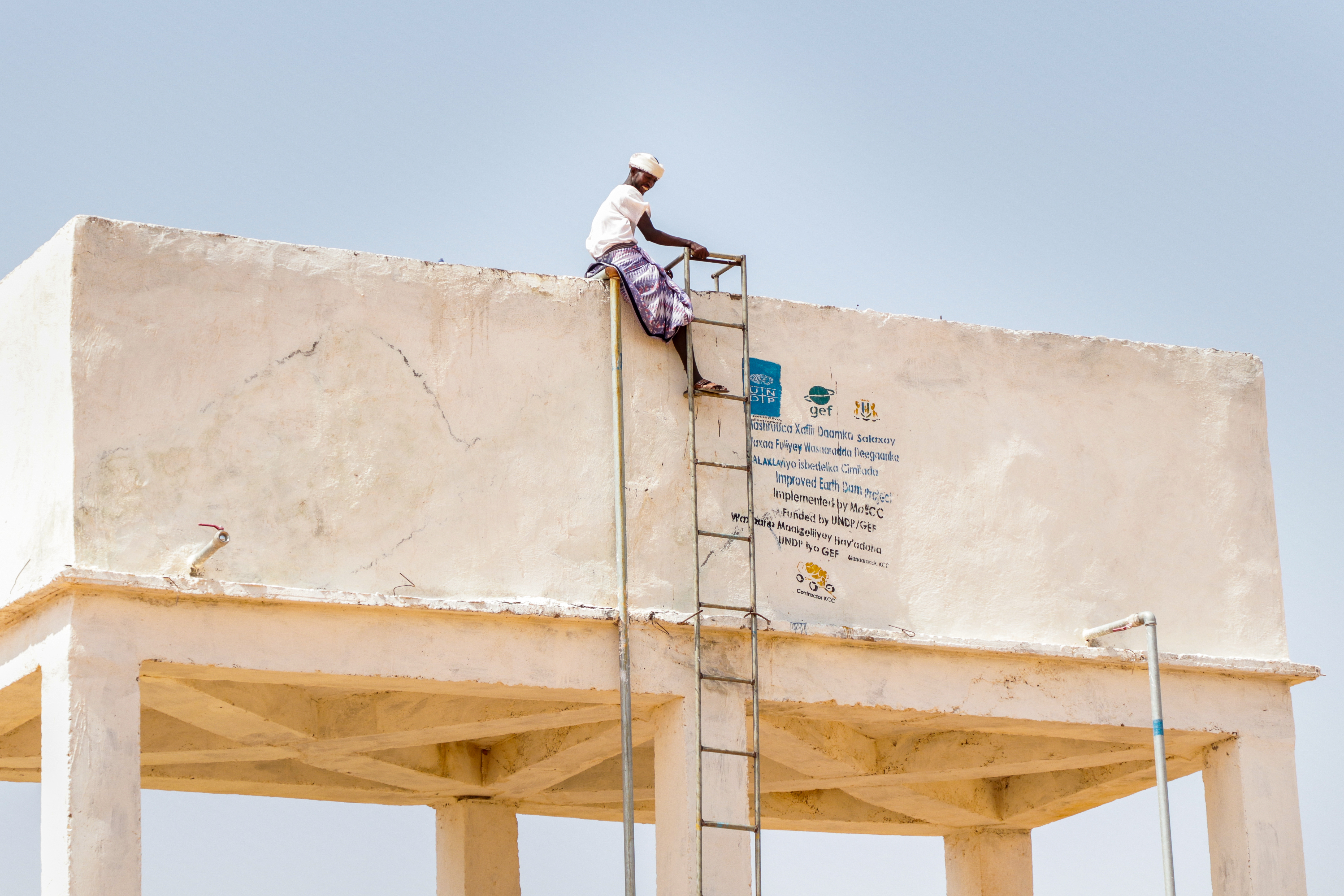 Ein Mann sitzt in Somalia oberhalb einer Leiter auf einem grossen steinigen Wasserbehälter, der auf Stelzen steht. Auf dem Behälter ist unteranderem ein UNDP-Logo